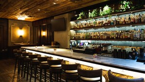A bar area with a long counter, multiple stools, and shelves stocked with bottles. - The Gotham Hotel (New York)
