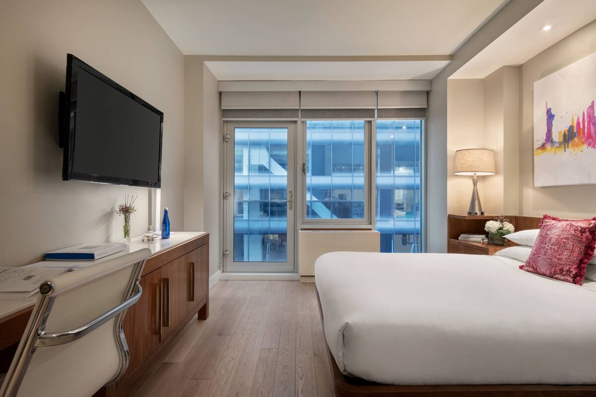 A modern hotel room with a large bed, a flat-screen TV mounted on the wall, a desk with a computer, and a view of the city through floor-to-ceiling windows.