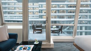 A modern hotel room with a large sliding glass door leading to a balcony with seating and a view of a building with glass windows. - The Gotham Hotel (New York)