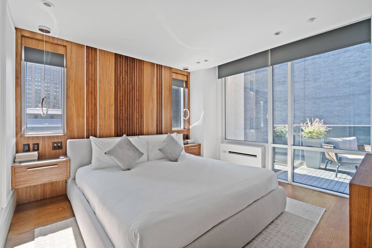 A modern bedroom with a large bed, wooden panel walls, a view of the city, and a balcony with outdoor furniture.