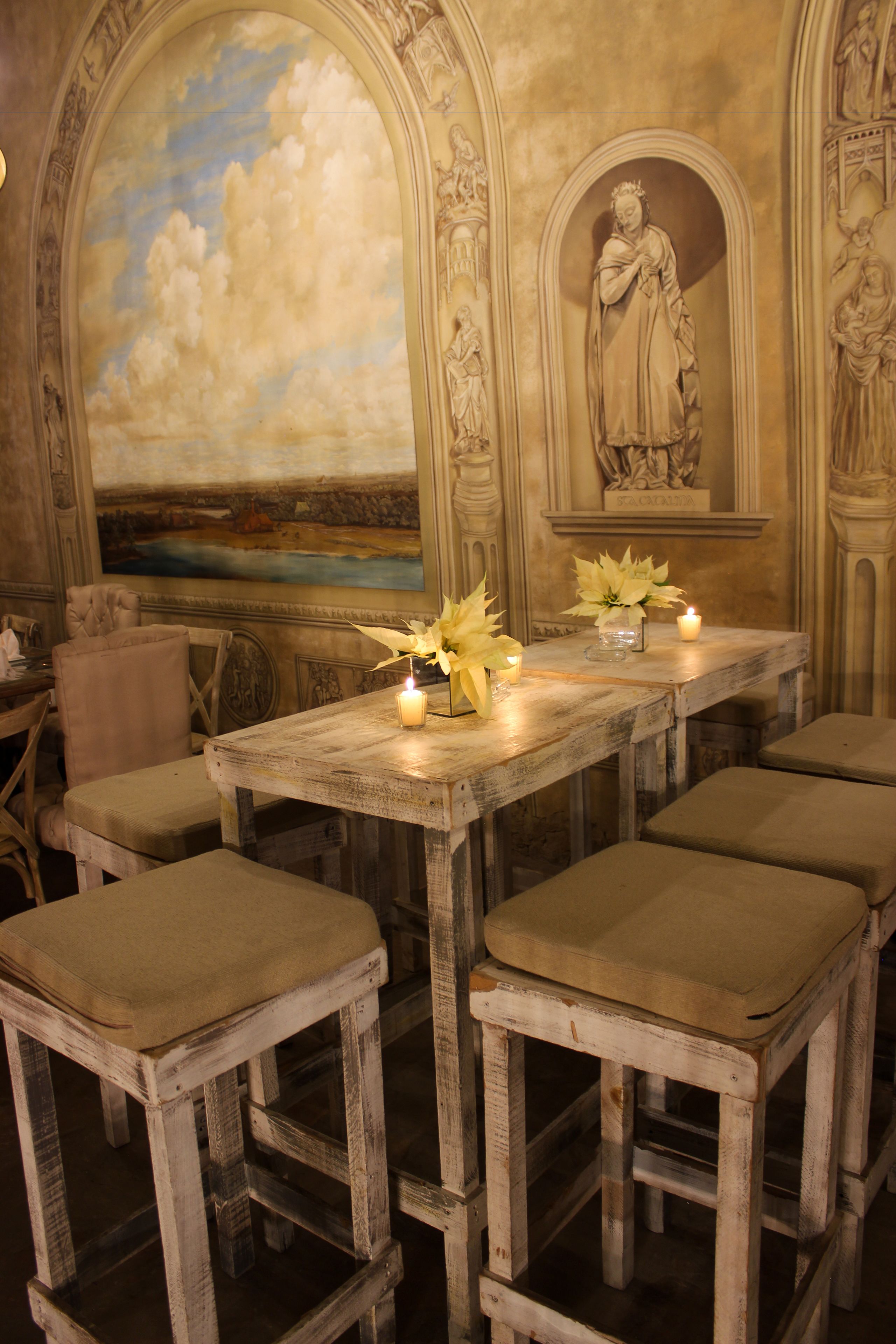 A dining area with a rustic wooden table, cushioned stools, and a mural of a landscape.