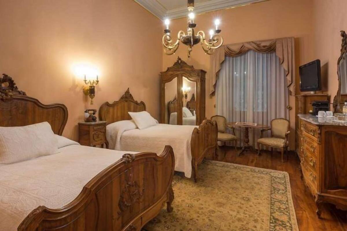 Une chambre d’hôtel avec deux lits en bois, un lustre, un miroir, une télévision et une petite table avec des chaises.