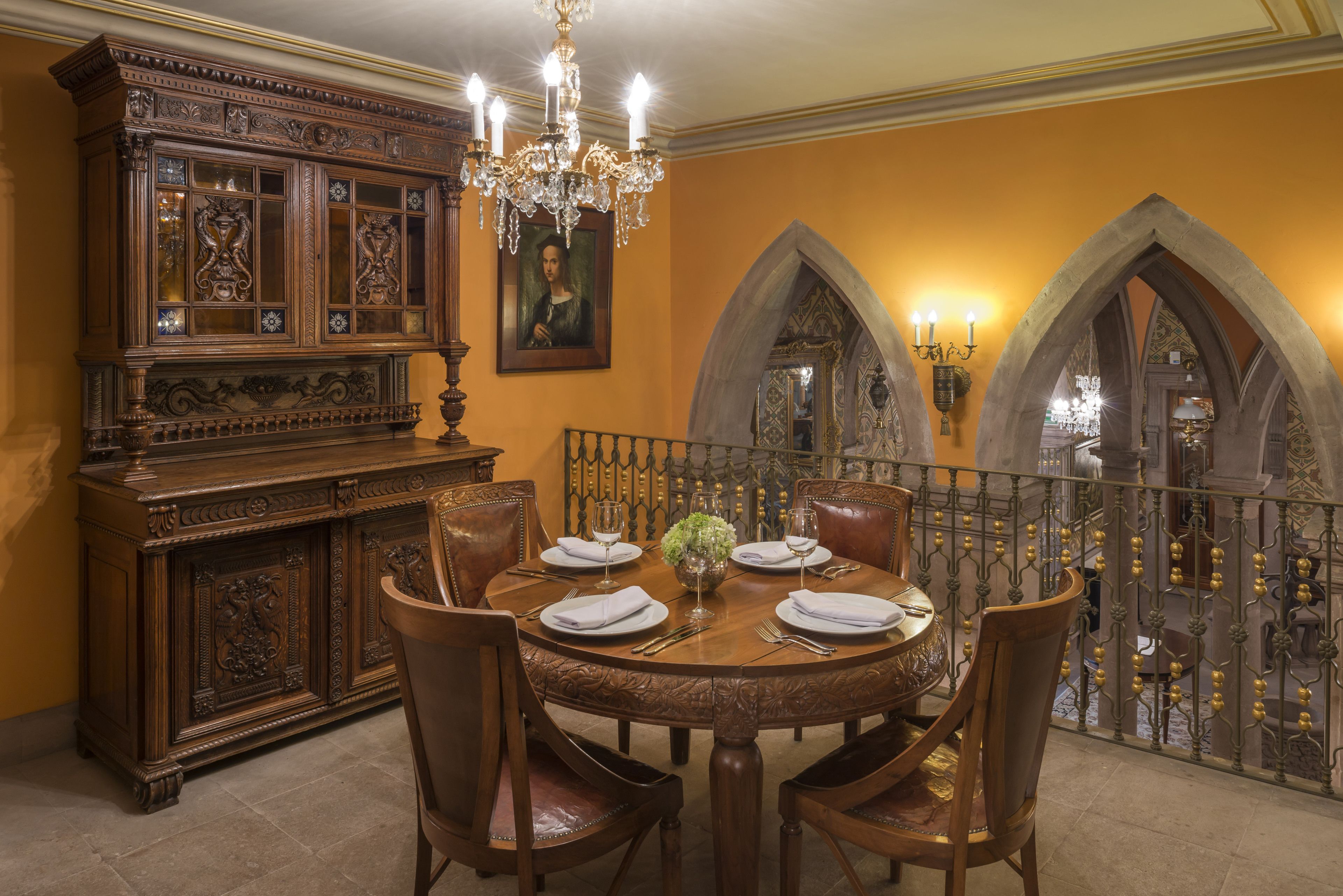 A dining area with a round wooden table set for four, surrounded by leather chairs. A large wooden cabinet with glass doors and intricate carvings is to the left. A chandelier hangs above the table, and a painting is on the wall. The room has arched doorways and a balcony with a wrought iron railing.