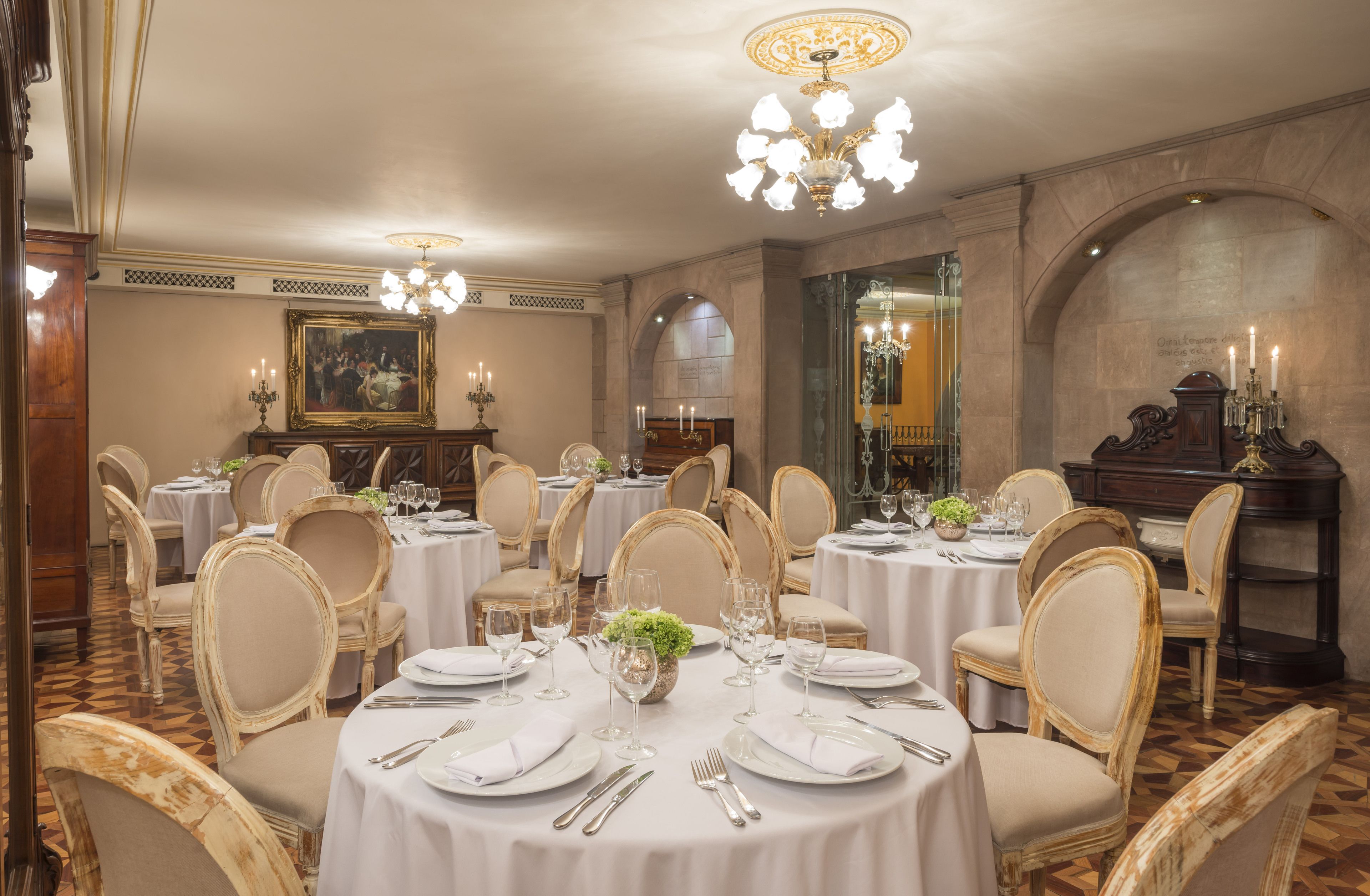 Un comedor formal con mesas redondas, manteles blancos y sillas con marcos dorados.