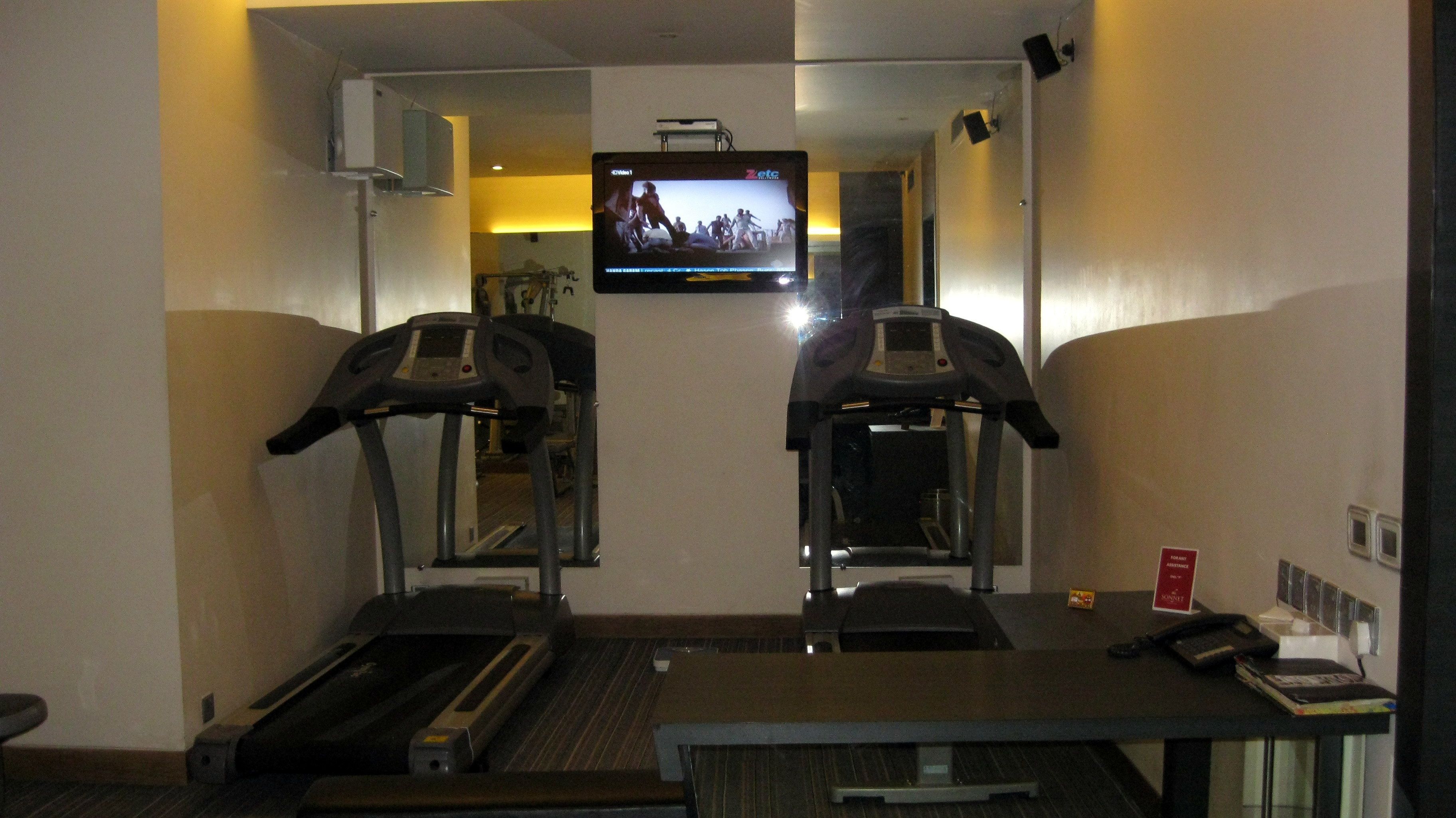 Habitación de hotel con dos cintas de correr, un televisor montado en la pared, una mesita con un teléfono y una revista, y un espejo que refleja la habitación.