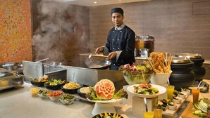 Montaje de bufé con diversos platos, un chef preparando comida y una cocina moderna al fondo.