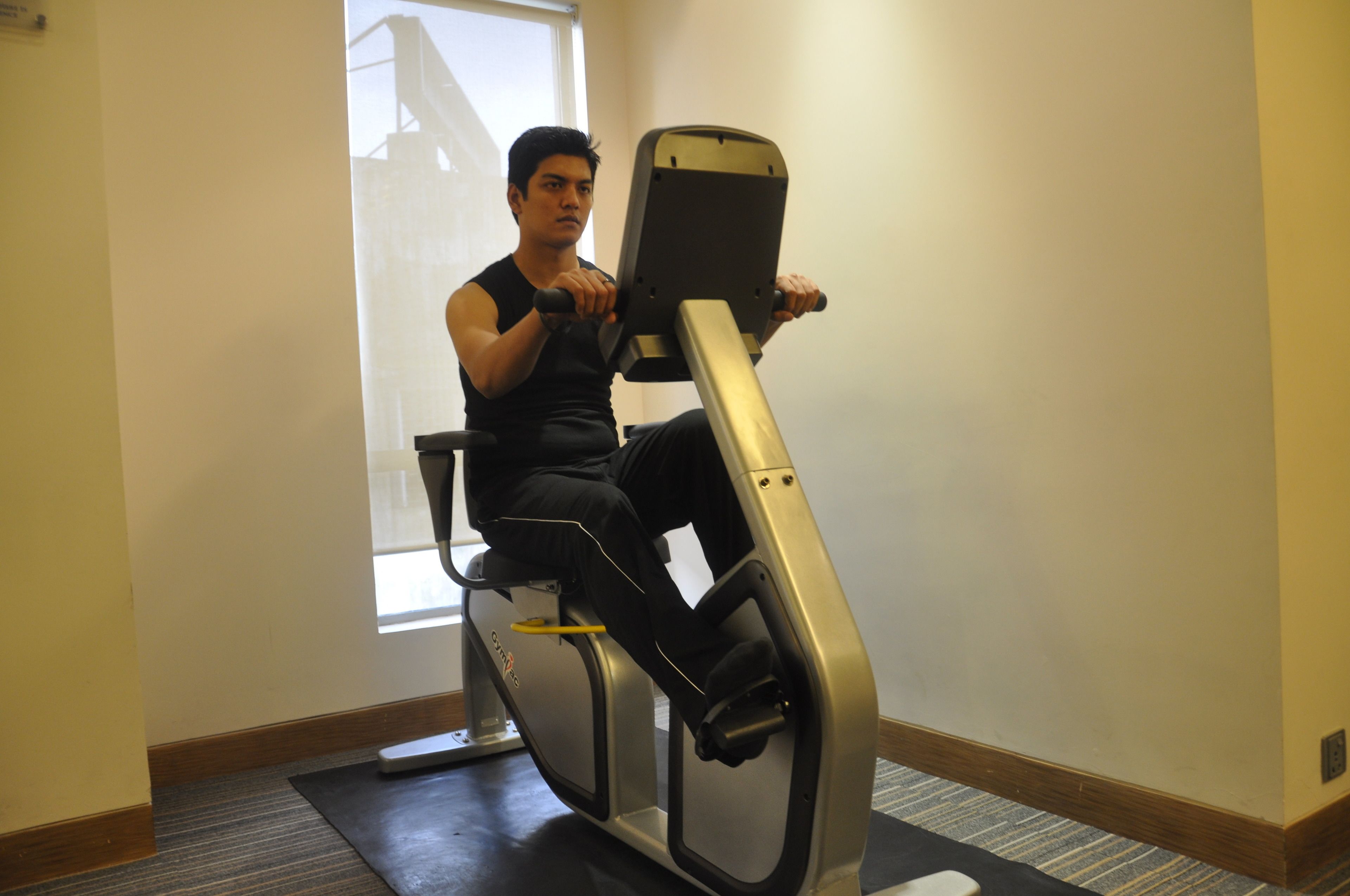 A person using a stationary bike in a room with a window, carpeted floor, and a plain wall.