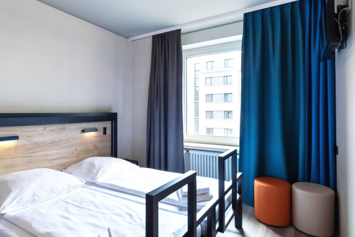 Ein Hotelzimmer mit einem hölzernen Kopfteil, einem Bett mit weißen Leinen, einem Fenster mit blauen Vorhängen und zwei orangefarbenen, zylindrischen Objekten auf der Fensterbank.