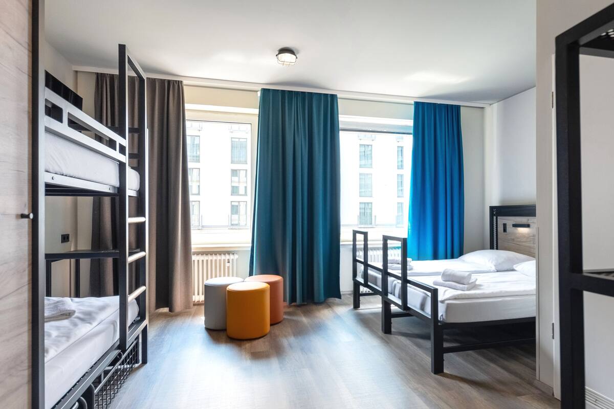 Ein Hotelzimmer mit Etagenbett, einem Einzelbett, einem kleinen orangefarbenen Hocker und Blick auf ein Gebäude durch große Fenster.