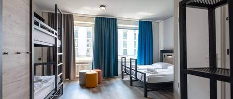 A hotel room with bunk beds, a single bed, a small stool, and a view of a building through a window with blue curtains.
