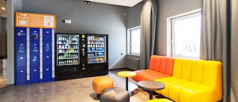 A modern lounge area with a yellow sofa, orange and gray ottomans, a vending machine, and a blue locker area.