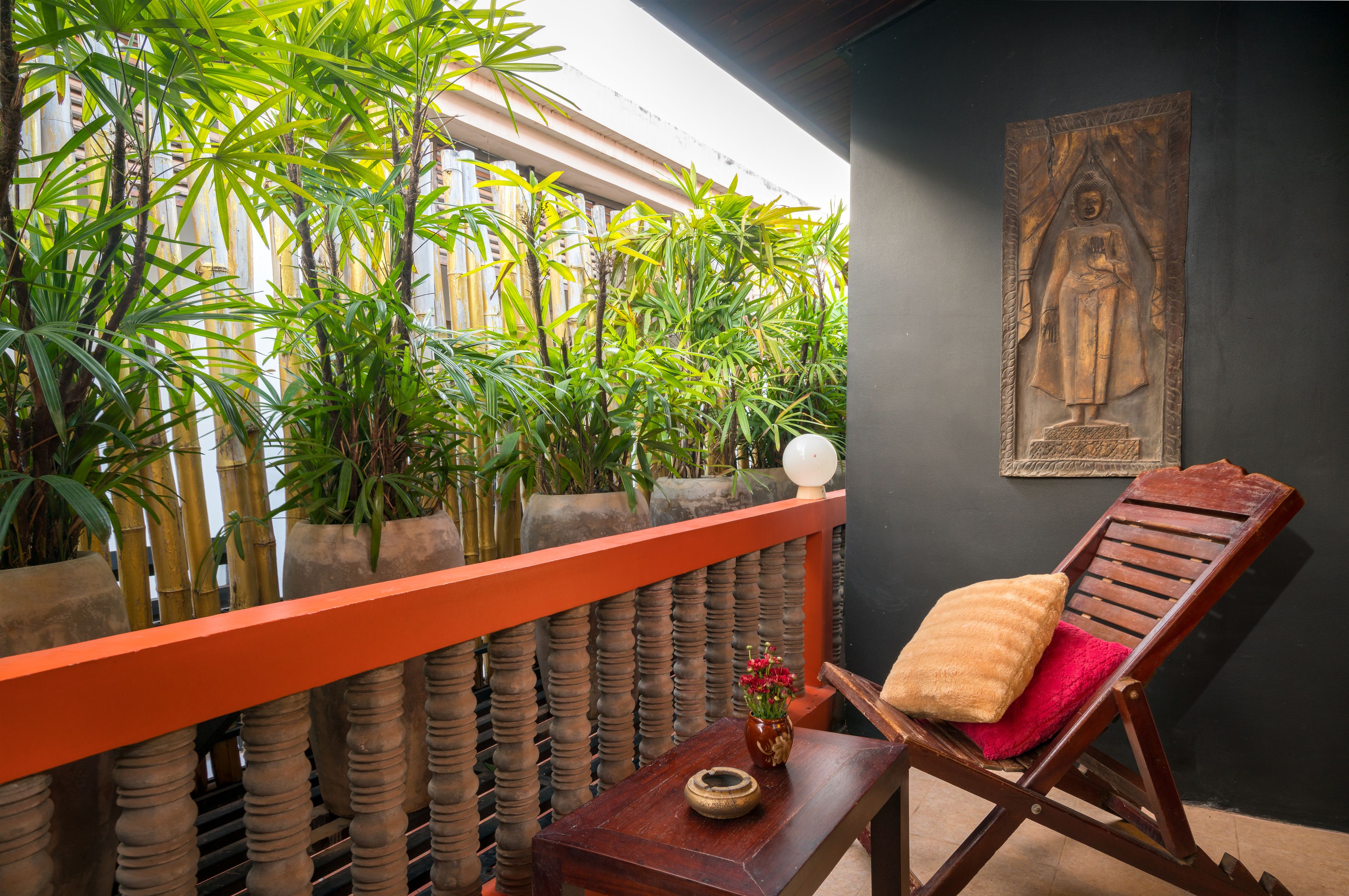 Un balcón con una silla de madera, una mesita con un florero y una estatua de Buda en la pared.