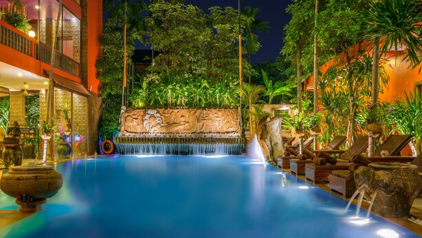 A pool area with a waterfall feature, surrounded by lush greenery and a stone wall with carvings.