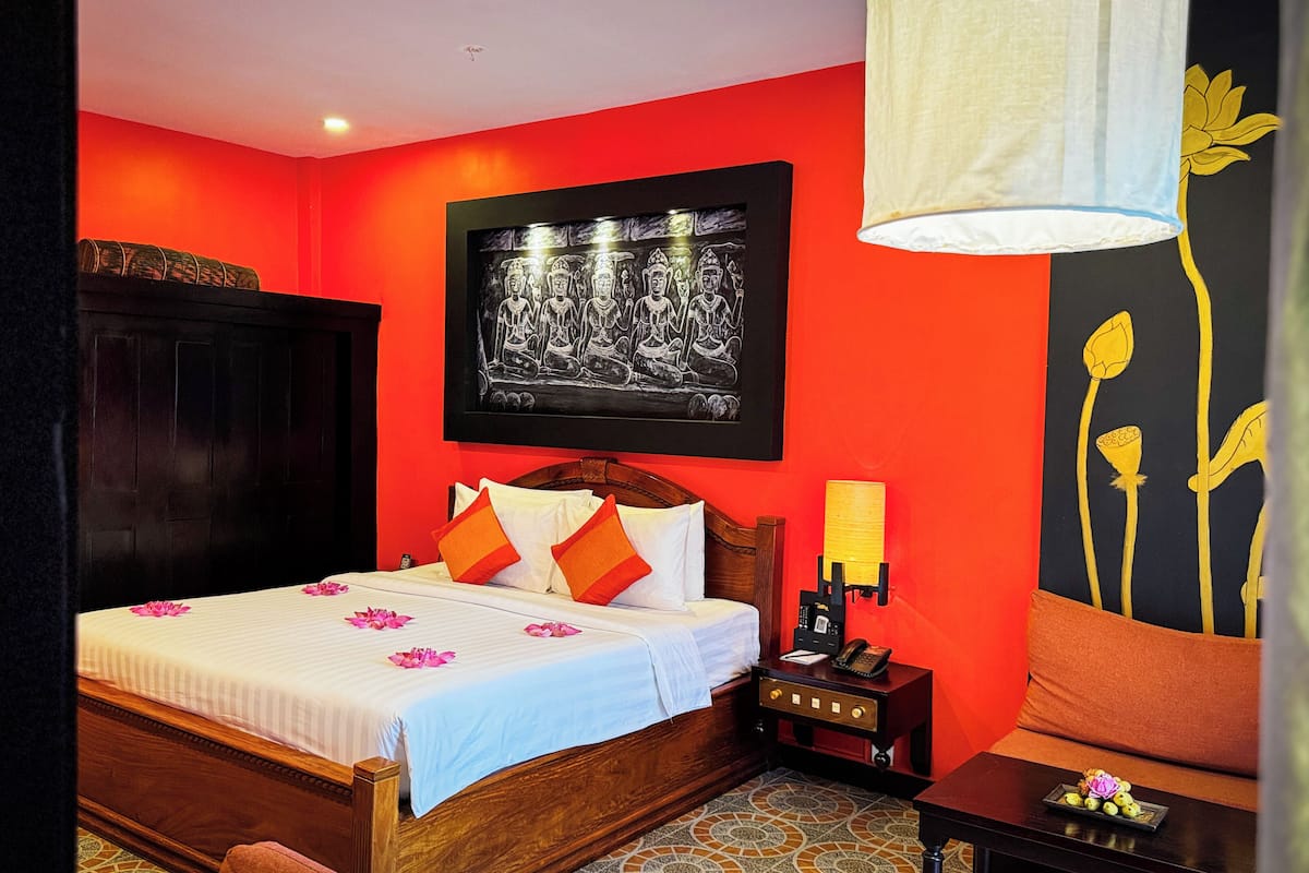 A hotel room with a wooden bed, red walls, a black and white painting, and a nightstand with a lamp.