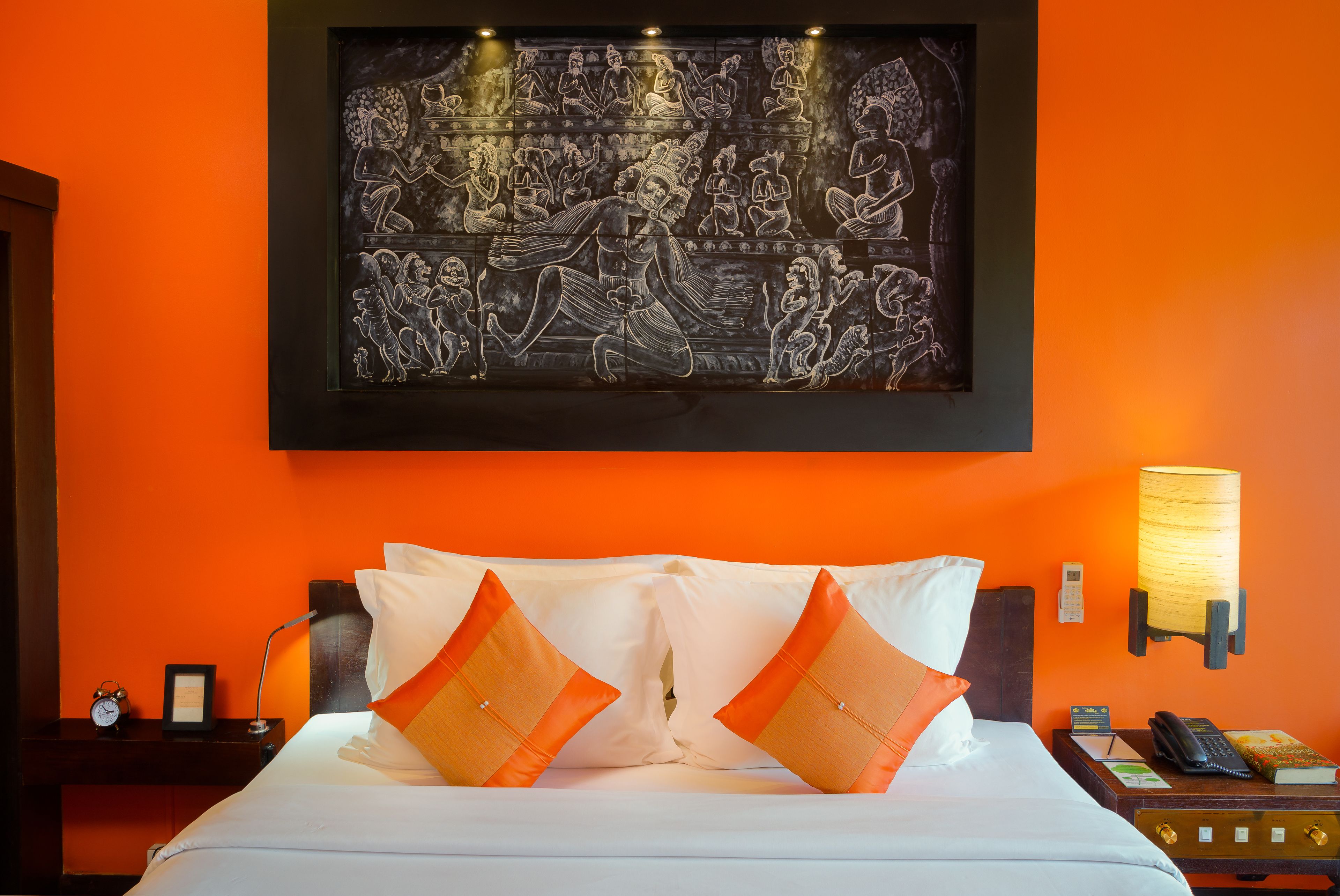 A hotel room with an orange wall, a bed with white and orange pillows, a framed artwork above the bed, a bedside lamp, a nightstand with a phone, and a lamp.