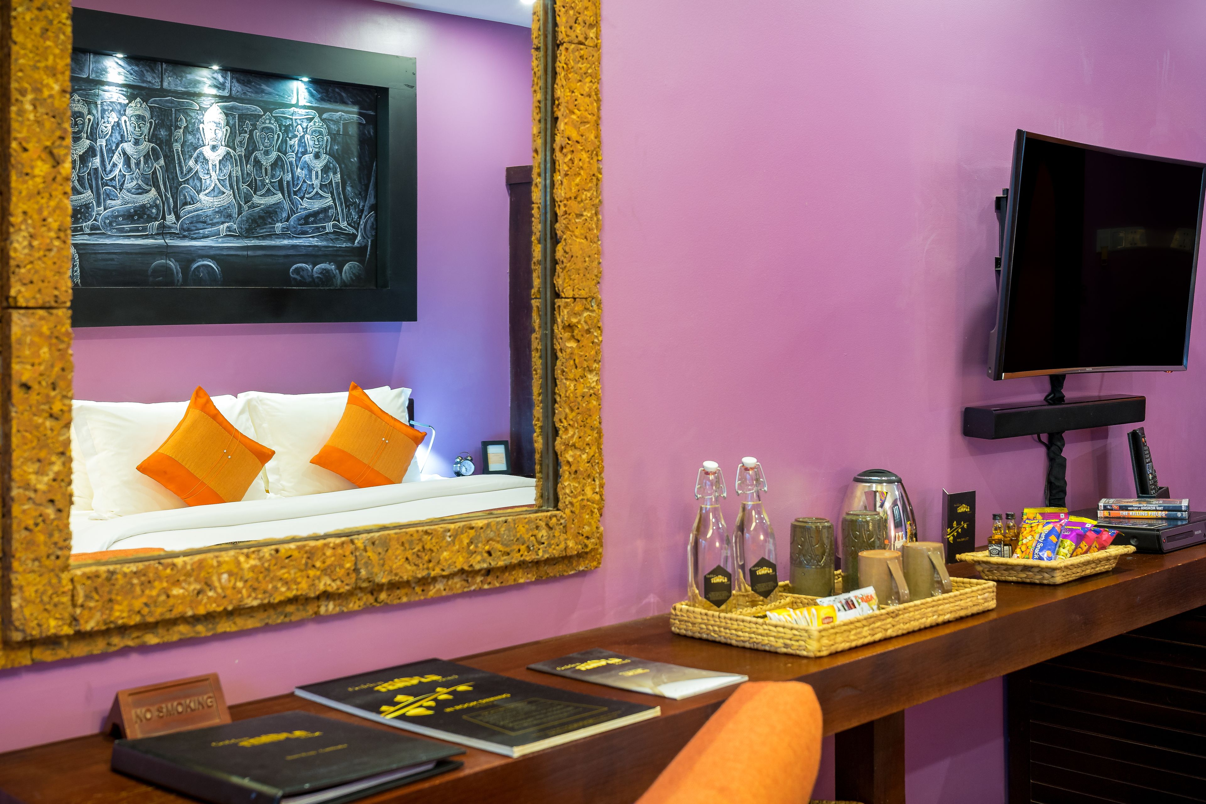 A room with a large mirror, a bed with orange pillows, a TV, and a reception desk with various items.