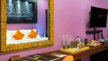 A room with a large mirror, a bed with orange pillows, a TV, and a reception desk with various items.