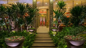 A hotel lobby with a central pathway leading to a reception area, surrounded by lush greenery and decorative planters.
