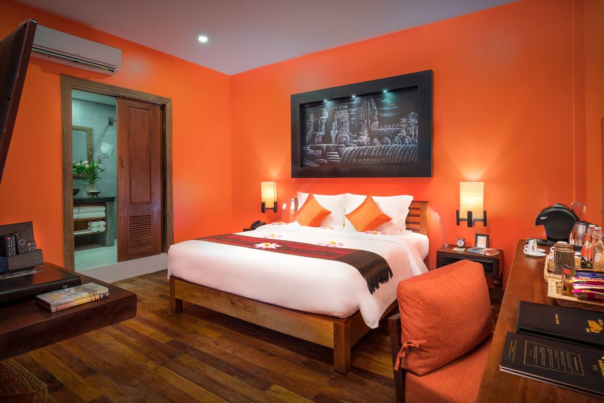 A hotel room with a large bed, wooden flooring, a desk with a computer and books, a chair, and a nightstand with a lamp and a clock.