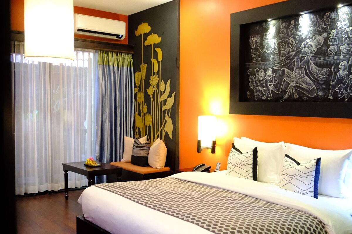 A hotel room with a large bed, a nightstand, a lamp, an air conditioner, and a wall with a floral design.