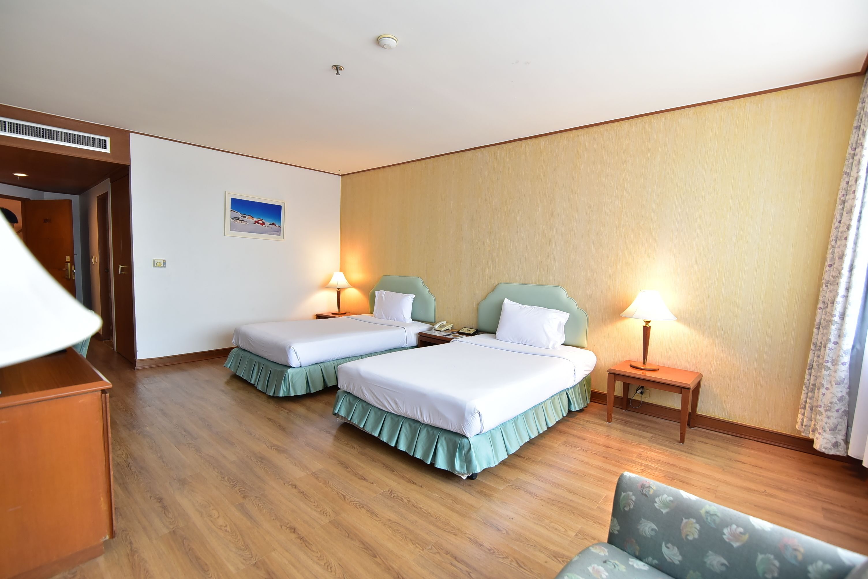 A hotel room with two beds, a wooden floor, a small table, a lamp, and a picture on the wall.