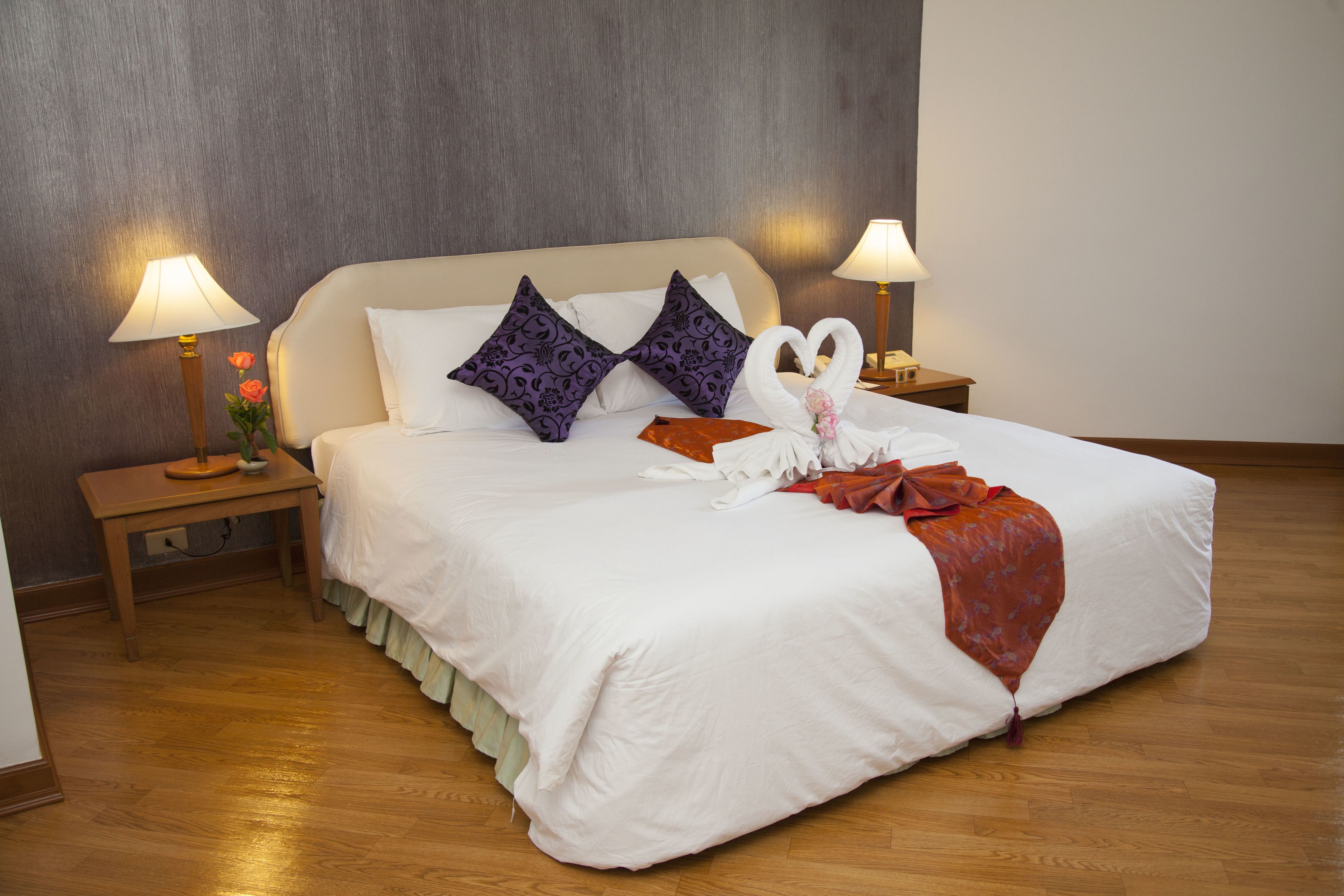 A neatly made bed with white linens, two purple patterned pillows, and a swan-shaped pillow. There are two bedside tables with lamps and a vase of flowers.