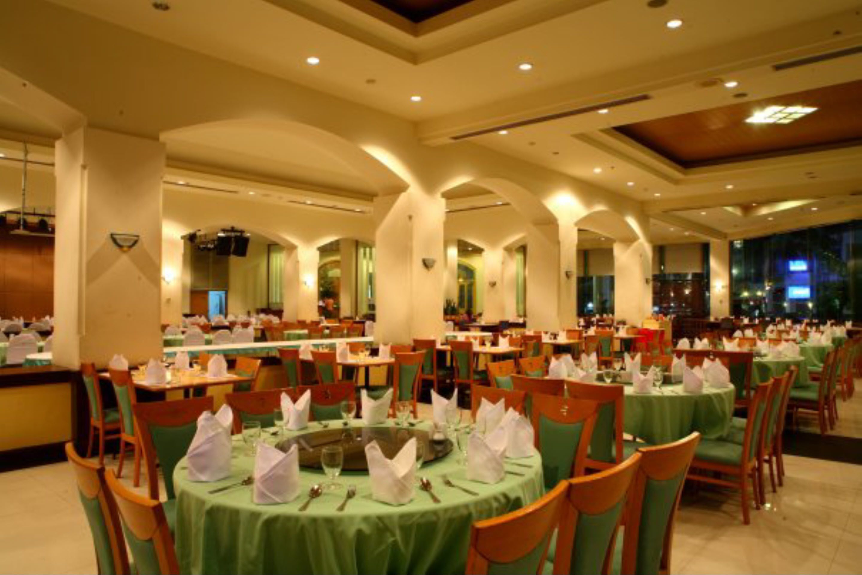 Un amplio comedor con mesas redondas cubiertas con manteles verdes y servilletas blancas, dispuesto en un espacio generoso con techos altos y columnas arqueadas.
