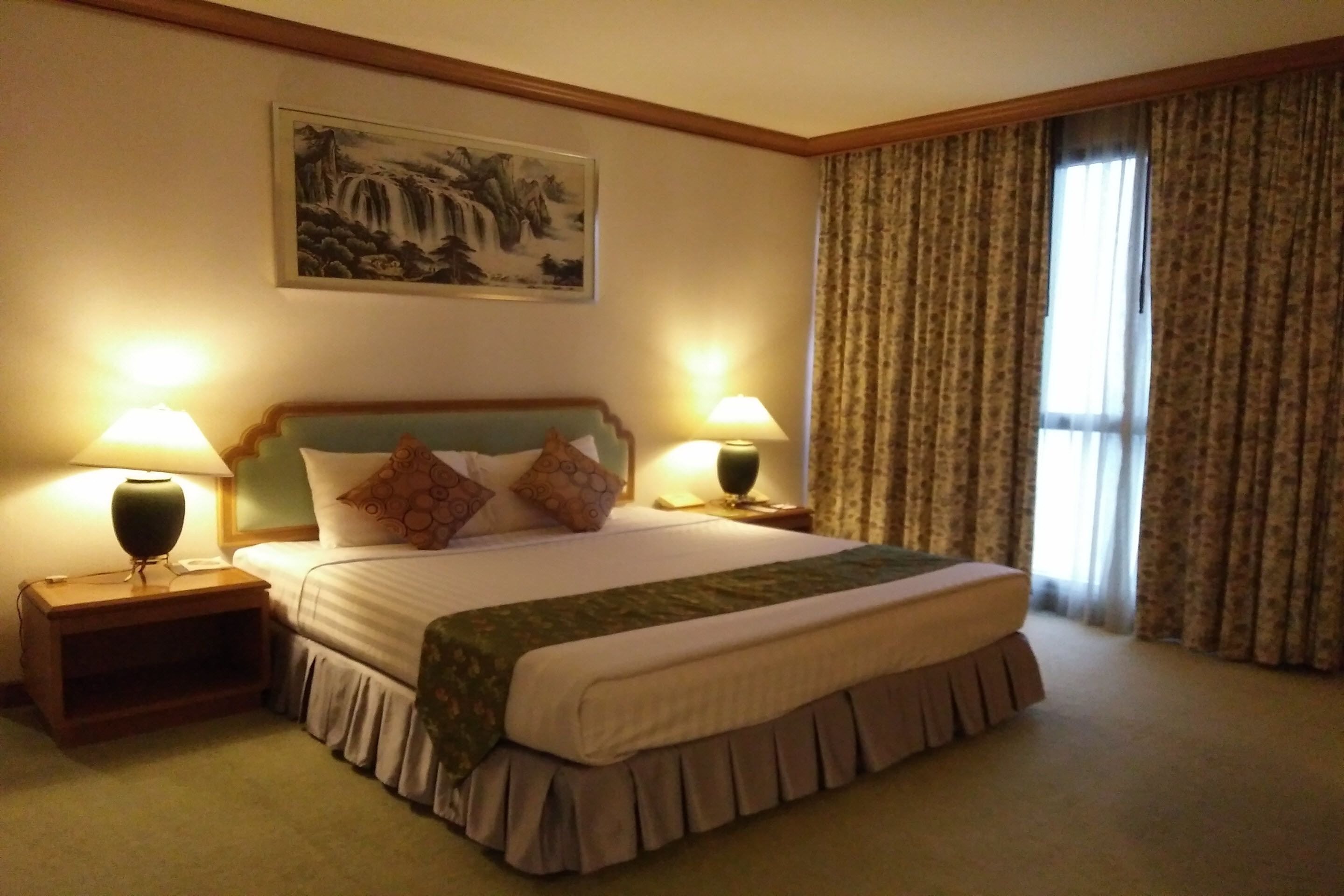 A hotel room with a large bed, two bedside lamps, a painting on the wall, and a window with curtains.