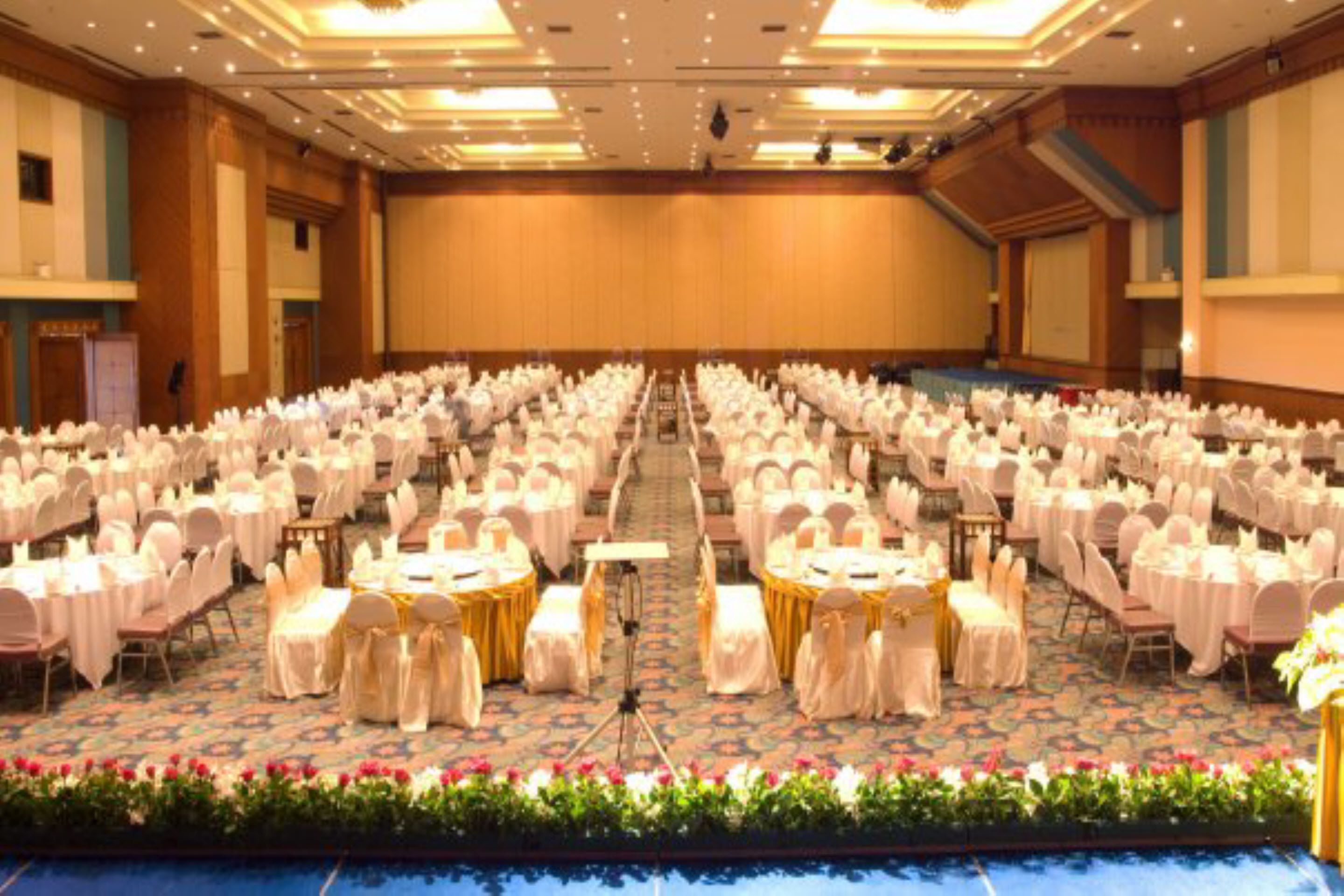 Un amplio salón de conferencias con filas de mesas redondas, cada una con mantel blanco y fajín dorado, y sillas con cubresillas blancos.