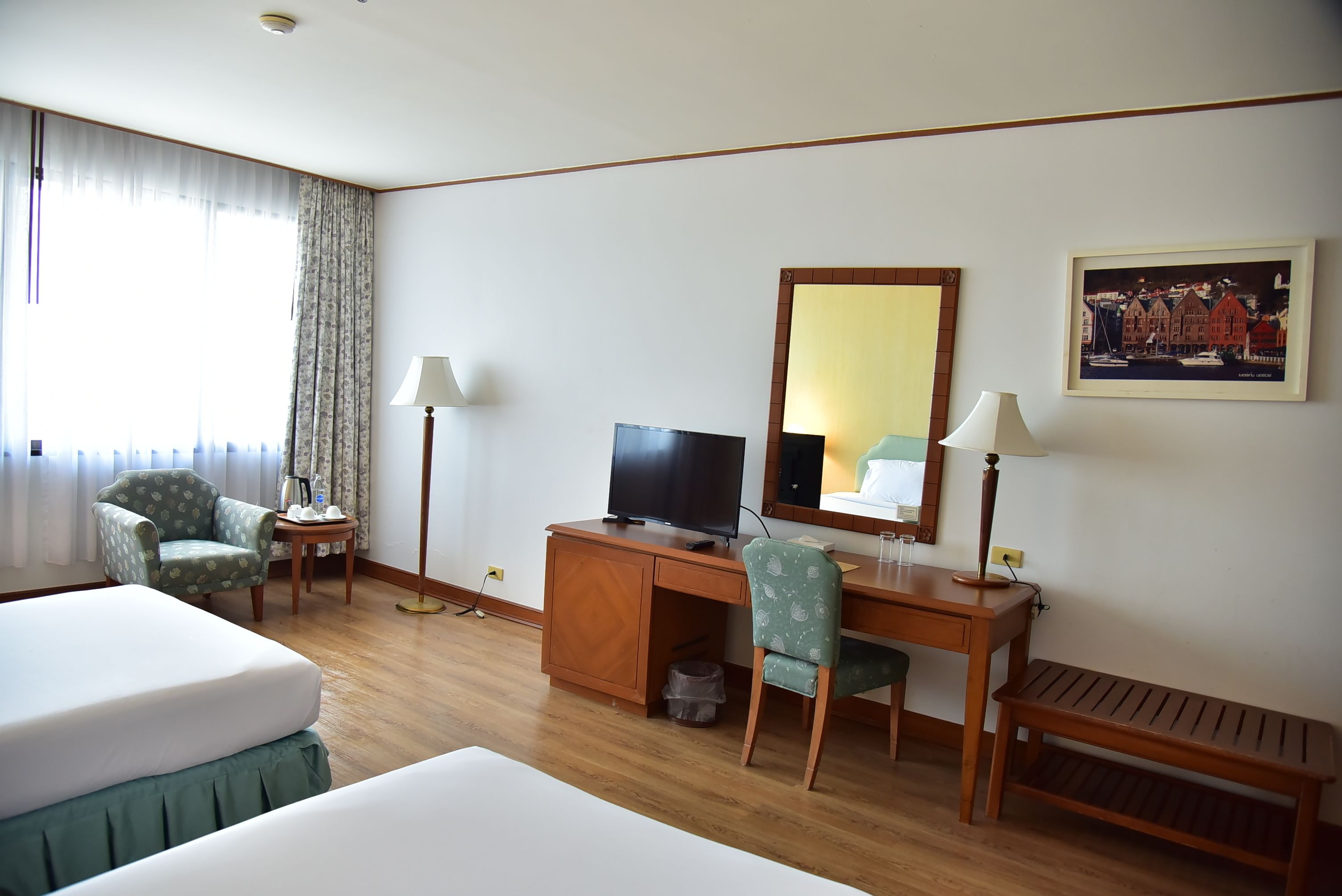 A hotel room with a bed, desk, chair, TV, lamps, and a painting on the wall.