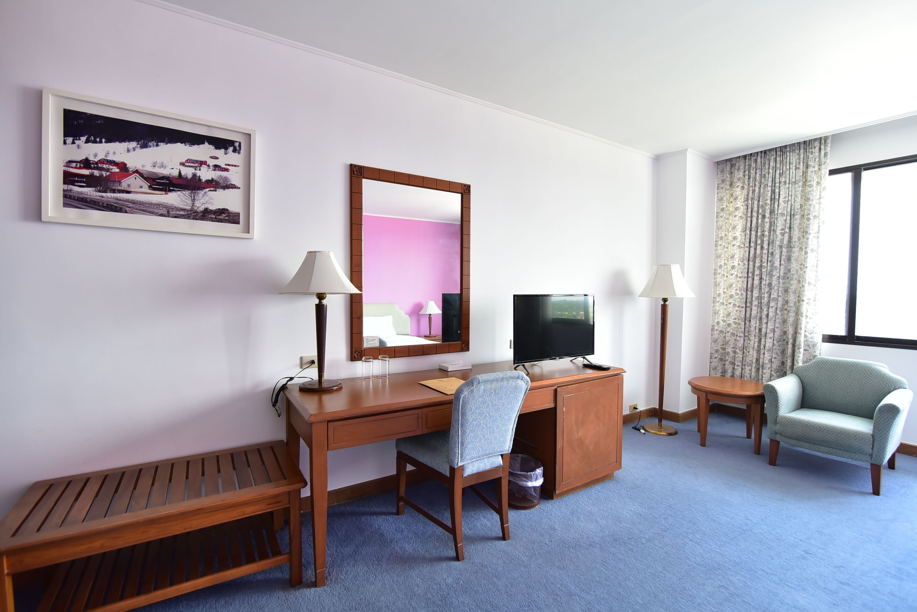 Une chambre d’hôtel avec un bureau en bois, une chaise, une télévision, un miroir, un banc et une fenêtre avec des rideaux.