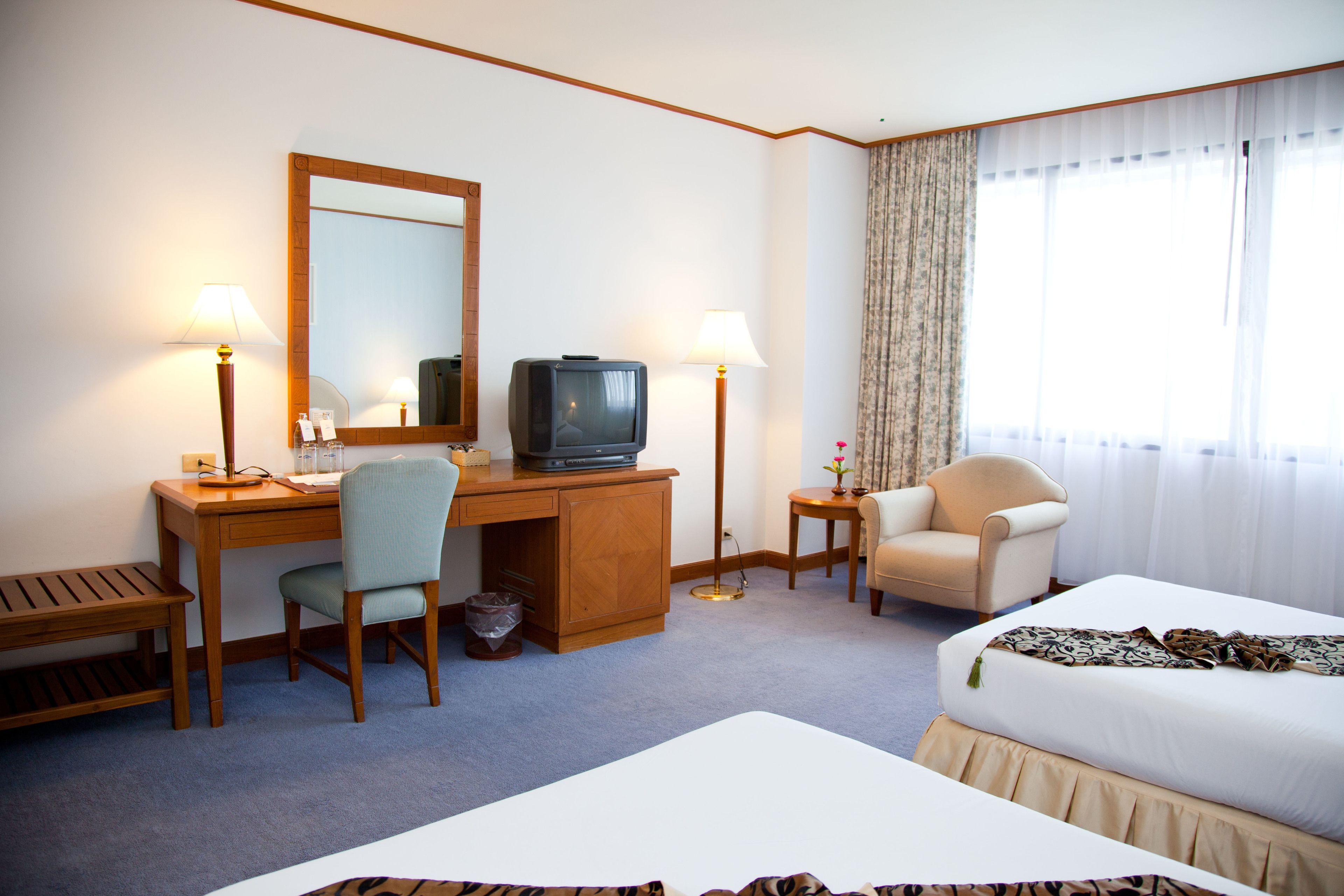 Habitación de hotel con cama, escritorio, silla, televisor, lámparas y un ventanal con cortinas.