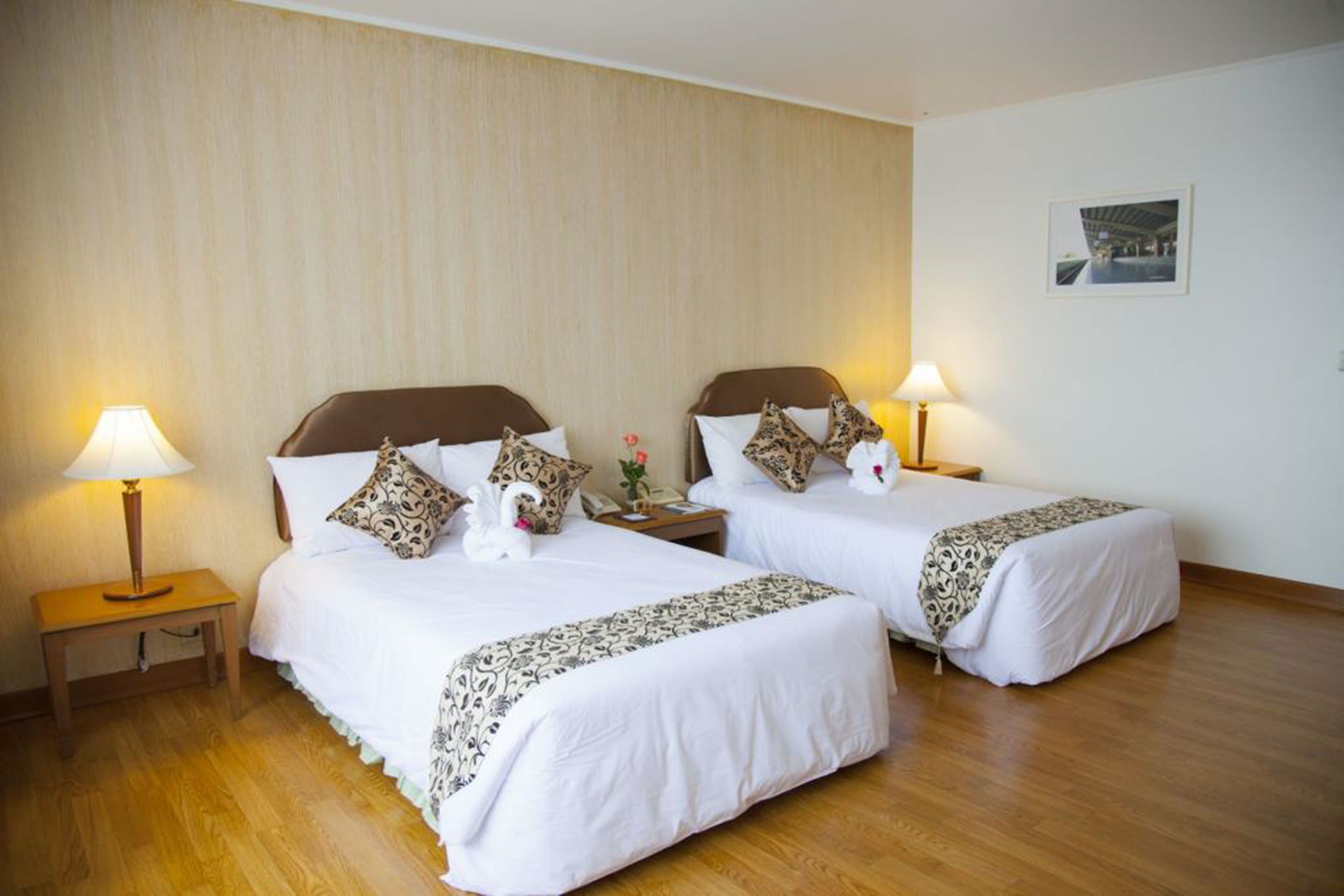 Habitación de hotel con dos camas, cada una con cabecero marrón, ropa de cama blanca y almohadas decorativas. Hay lámparas de noche y una mesita con lámpara entre las camas.