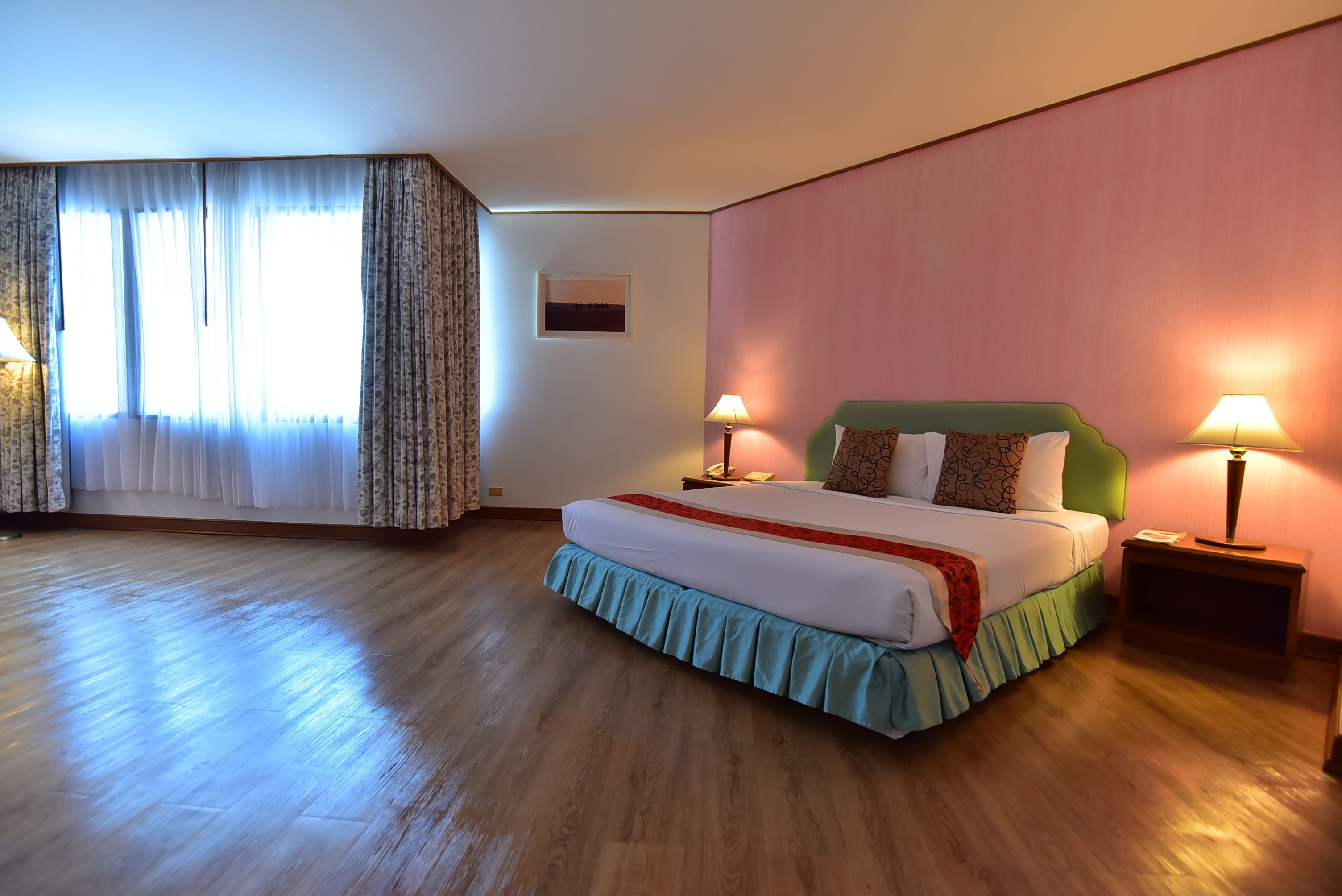 A hotel room with a large bed, two bedside lamps, a window with curtains, and a framed picture on the wall.