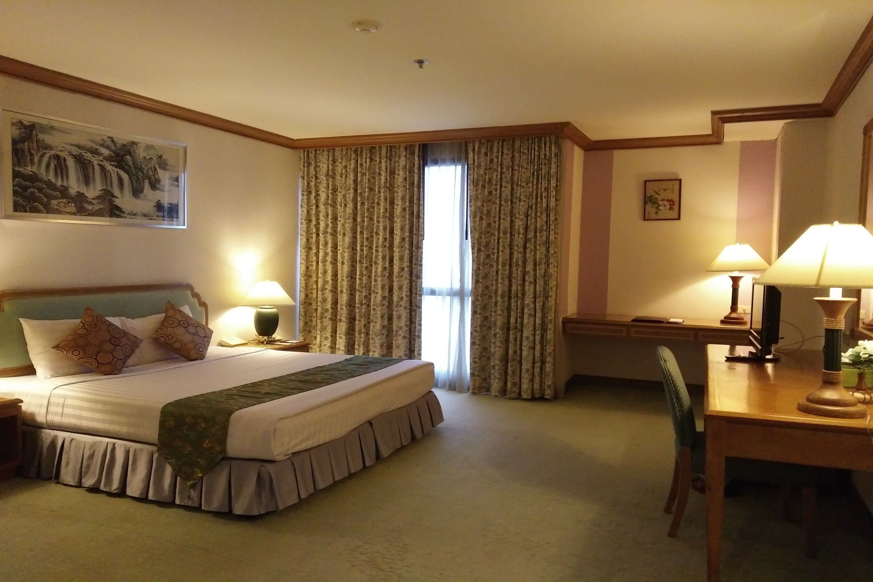 A hotel room with a large bed, two bedside lamps, a desk with a chair, and a window with curtains.