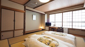 A traditional Japanese-style room with tatami flooring, two beds with floral bedding, a television, and a desk with a lamp. - Imari Grand Hotel (Imari)