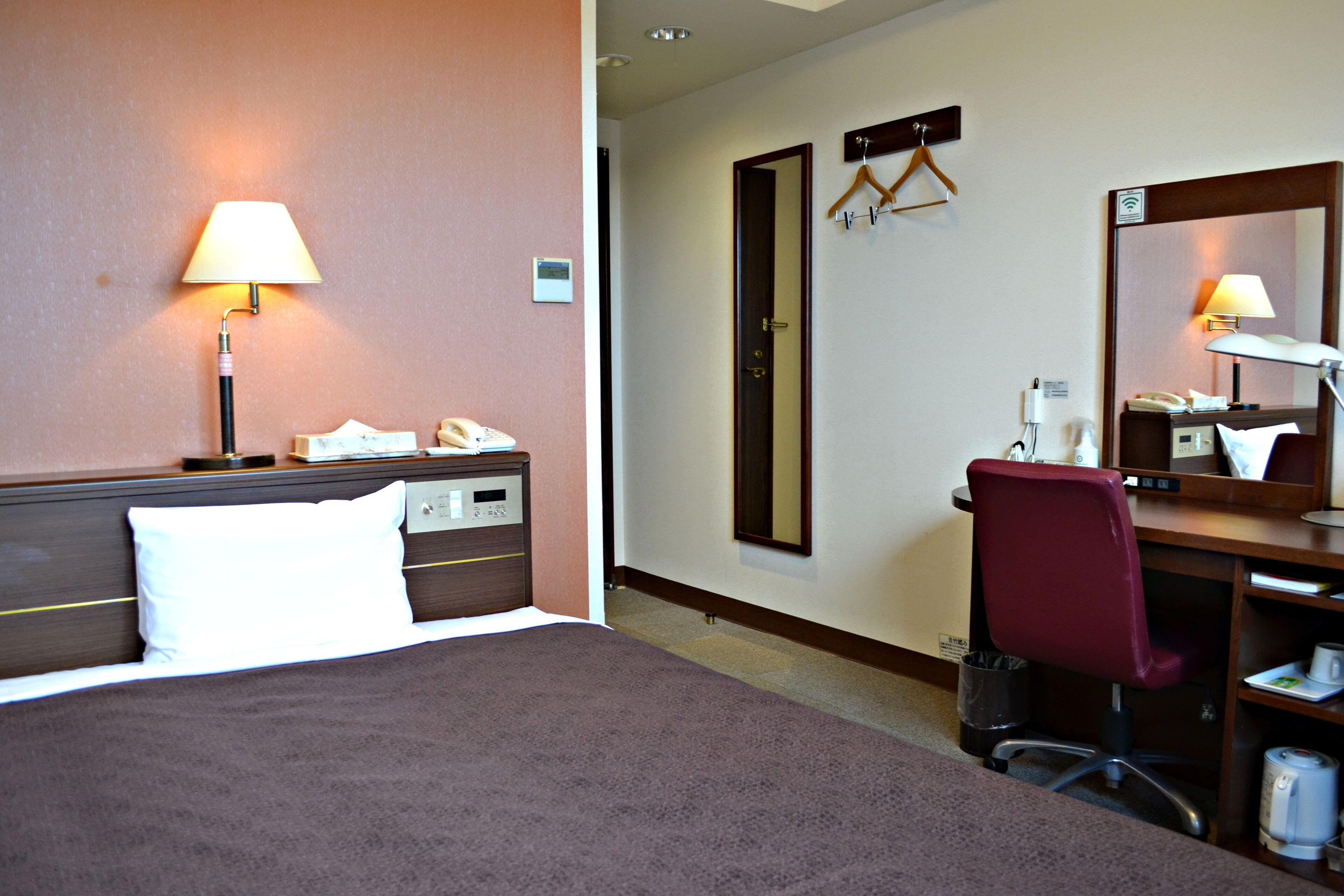 A hotel room with a bed, a desk, a chair, a mirror, and a lamp.