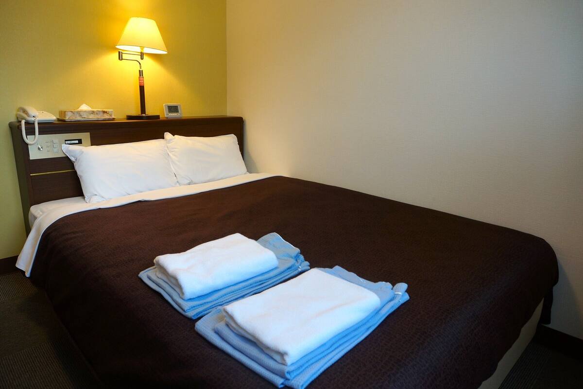 Habitación de hotel con una cama bien hecha, una lámpara de noche, un teléfono y un reloj digital en la mesita de noche.