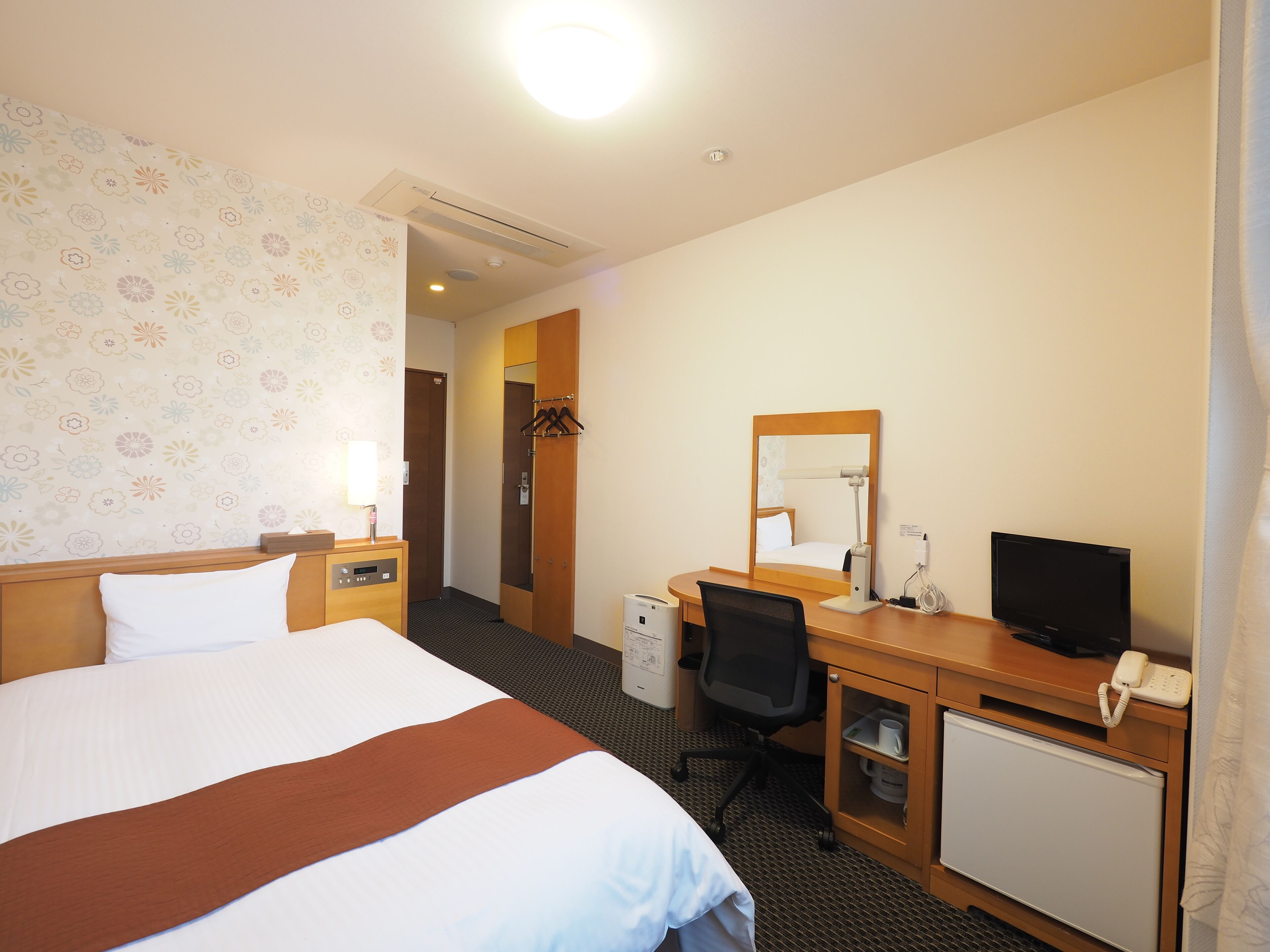 Quarto de hotel com cama, escrivaninha, cadeira, televisão e espelho.