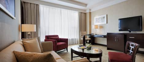 A hotel room with a sofa, armchair, coffee table, TV, and a desk with a lamp.