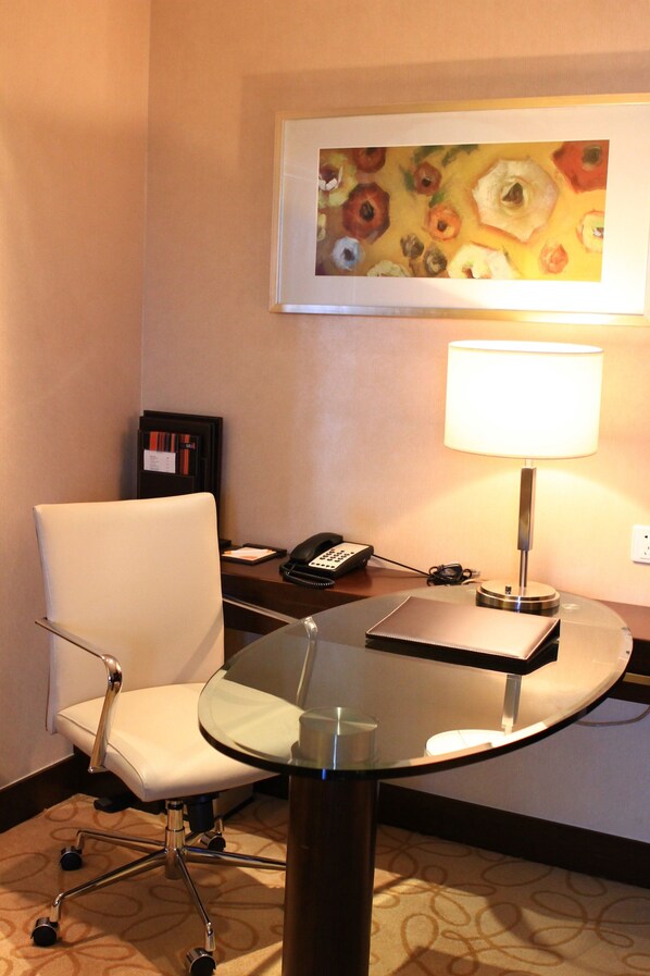 A hotel room with a glass table, a white chair, a desk with a lamp, a telephone, and a framed painting on the wall. - Courtyard by Marriott Hangzhou Wulin (Hangzhou)