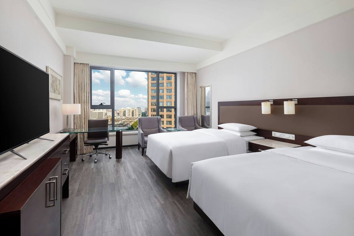 Une chambre d’hôtel avec deux lits, un bureau avec une télévision, une chaise et une grande fenêtre donnant sur la ville.