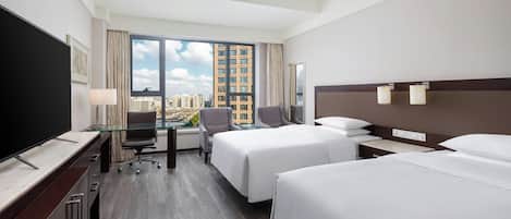 Hotelkamer met twee bedden, een bureau met een televisie, een stoel en een groot raam met uitzicht op de stad.