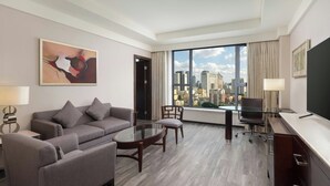Une chambre d’hôtel moderne avec un canapé gris, une table à manger en verre, un bureau avec une chaise et une grande fenêtre donnant sur la ville.