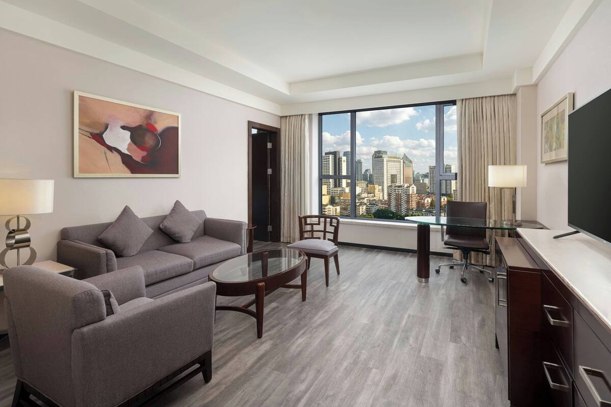 Une chambre d’hôtel moderne avec un canapé gris, une table à manger en verre, un bureau avec une chaise et une grande fenêtre donnant sur la ville.