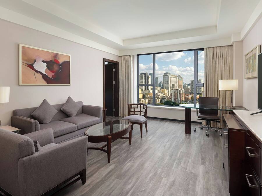 Una habitación de hotel moderna con un sofá gris, una mesa de comedor de cristal, un escritorio con una silla y un ventanal con vistas a la ciudad.