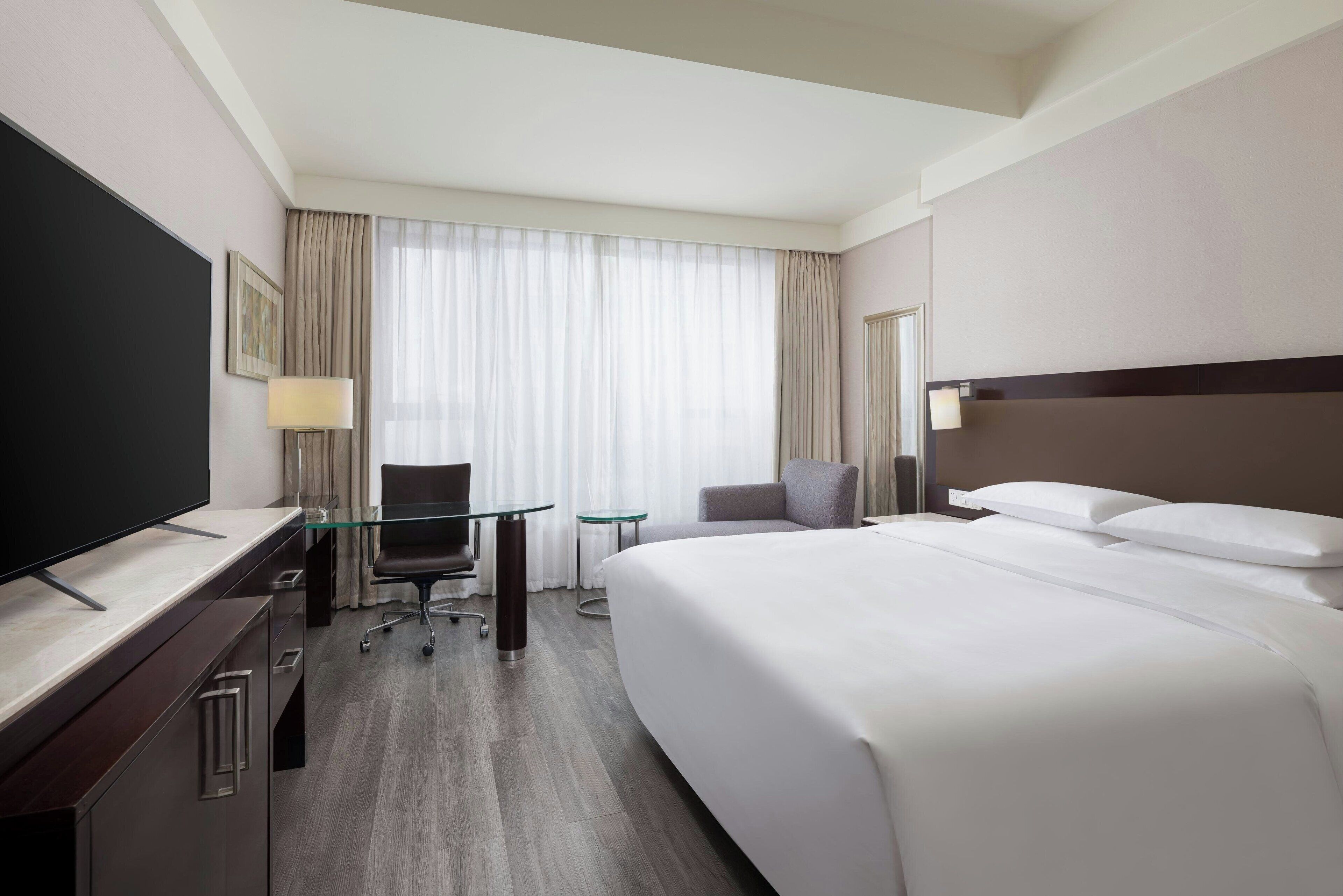 A hotel room with a large bed, a desk with a flat-screen TV, a chair, a small table, and a window with curtains.