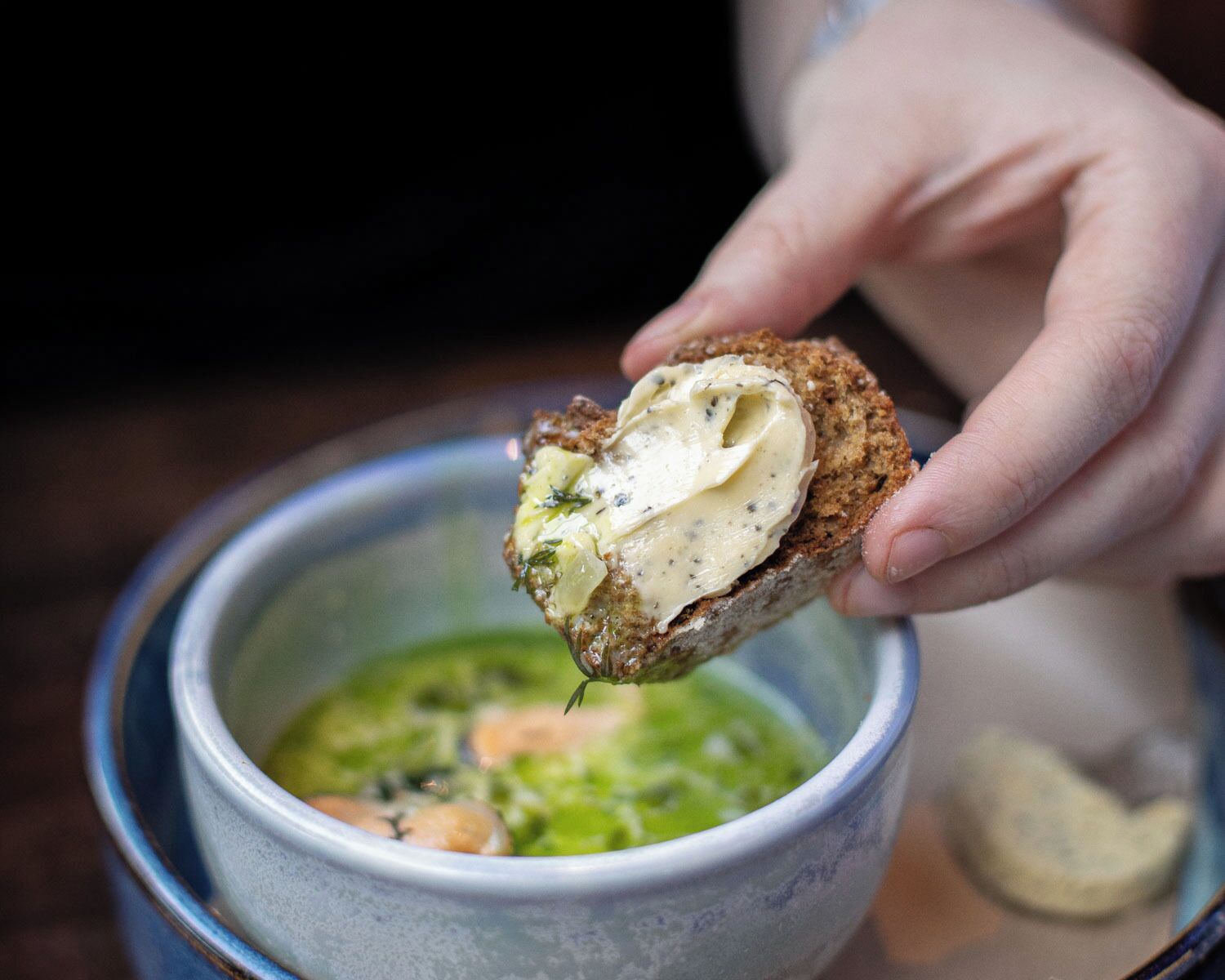 Eine Hand hält ein Stück Brot mit einer cremigen Aufstrich, über einer Schüssel grüne Suppe.