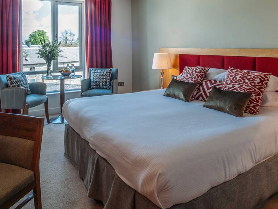 A hotel room with a large bed, two armchairs, a small table with a vase, and a view of the outdoors through a window with red curtains.