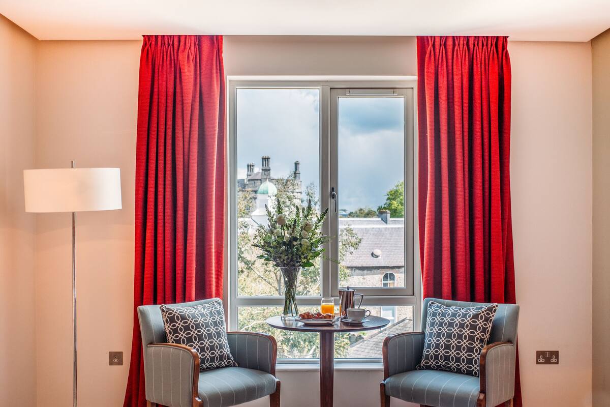 A hotel room with two armchairs, a table with breakfast items, a vase with flowers, and a view of buildings through a window with red curtains.